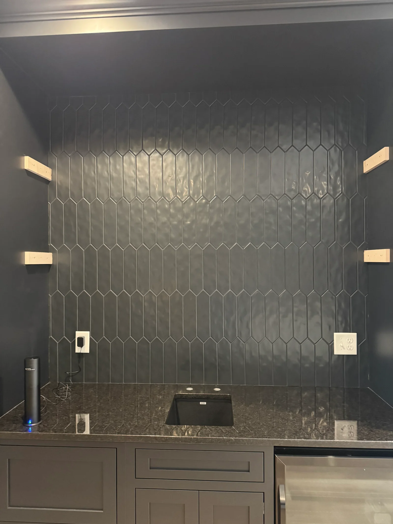 Gray hexagonal tile backsplash with recessed lighting and electrical outlets above granite countertop