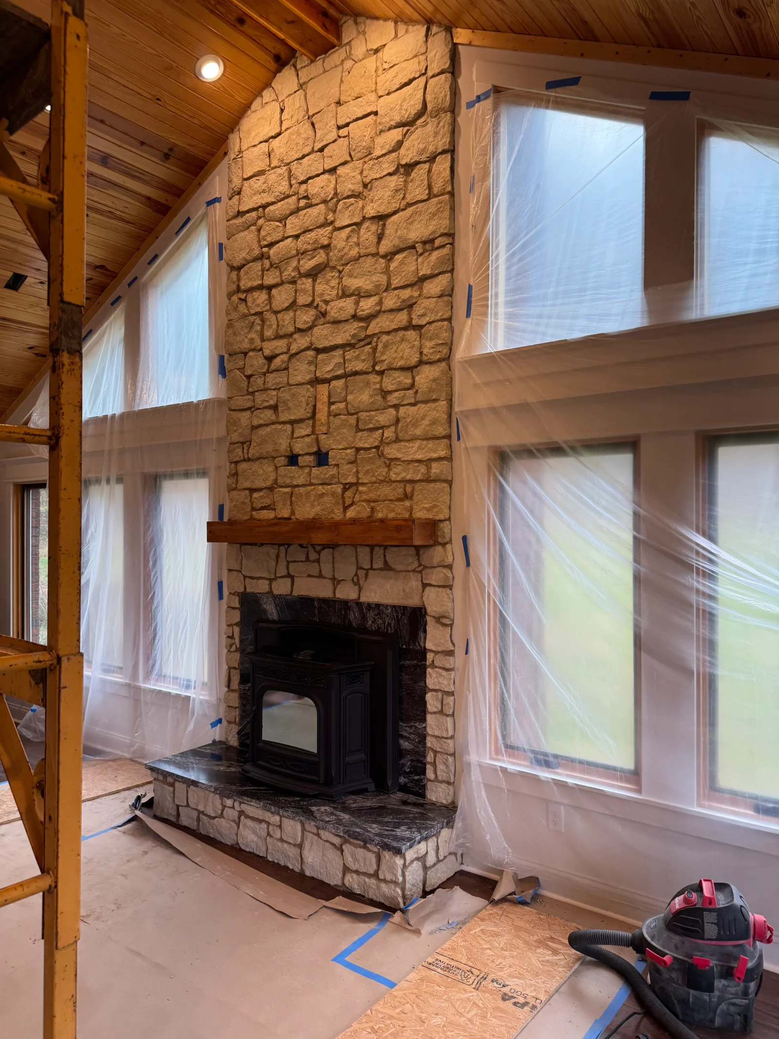 Tan stone fireplace with black stove insert, wooden mantel, and granite hearth during construction